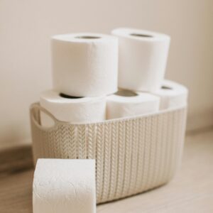 A woven basket filled with stacked white toilet paper rolls on a light wooden floor, perfect for hygiene and cleaning themes.