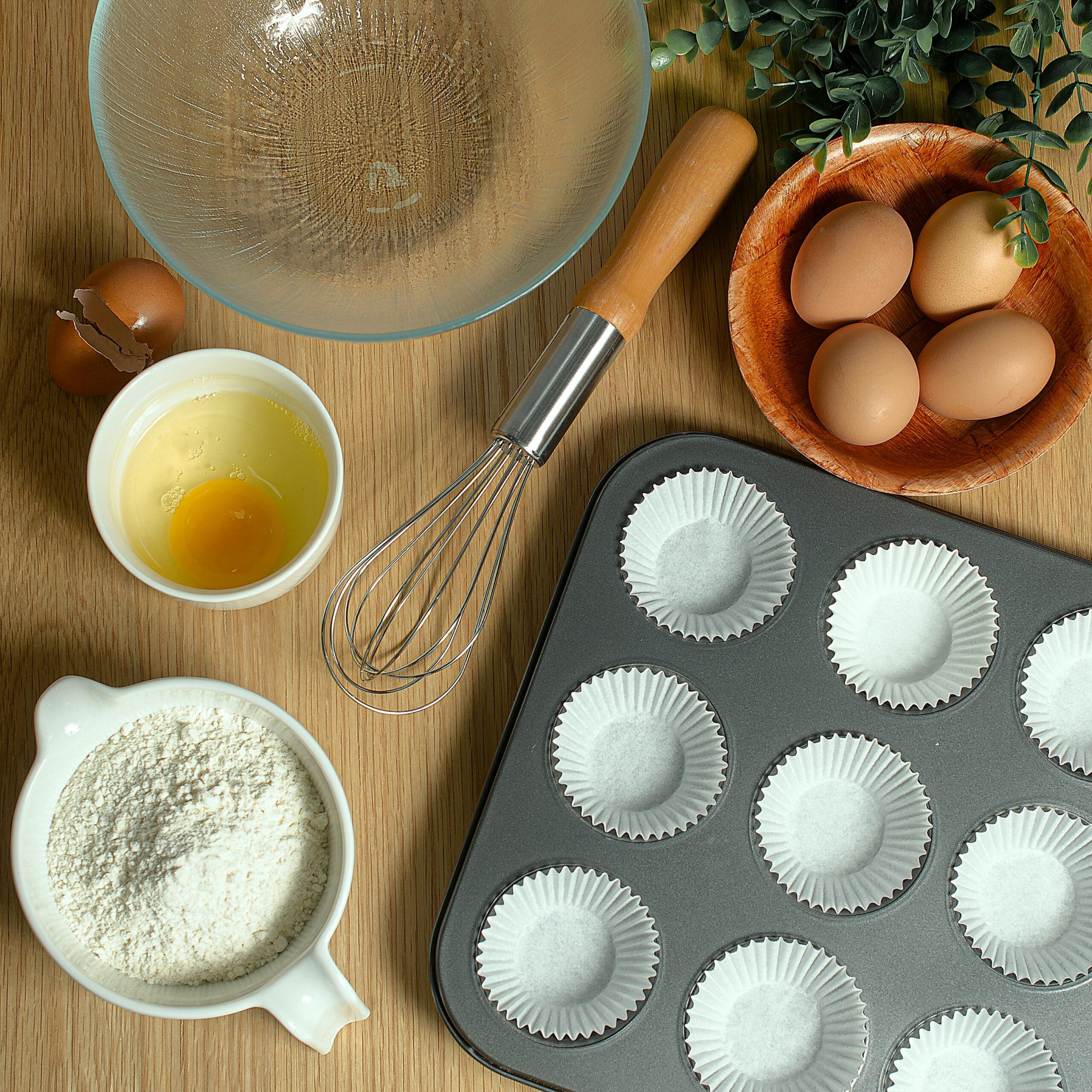 Flat lay of baking ingredients: eggs, flour, and a whisk on a wooden surface.